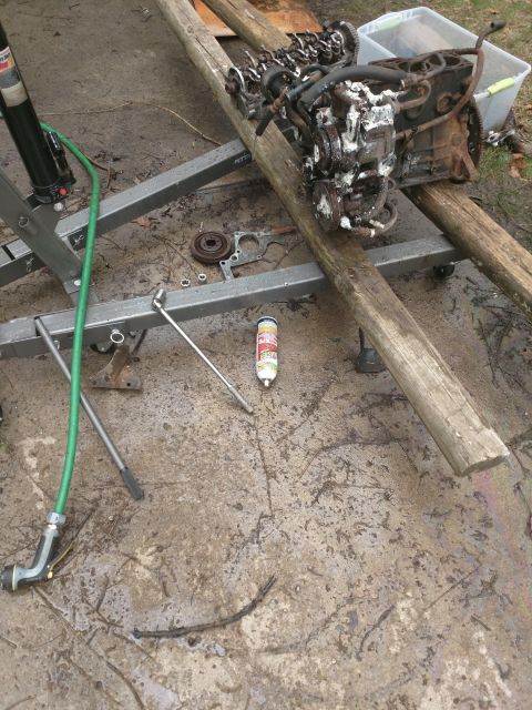1989 Ford Probe - An engine block sits on wooden beams supported by an engine hoist. Various tools and parts are scattered on the wet concrete ground around…