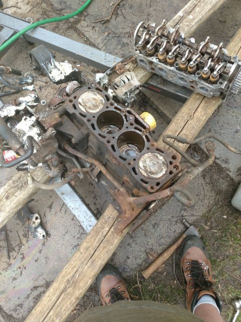 1989 Ford Probe - A disassembled engine block and cylinder head are laid out on wooden planks and a metal stand on a dirt and gravel surface. Various engine…
