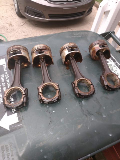 1989 Ford Probe - Four used pistons and connecting rods are laid out on a dark green surface, possibly a trash bin lid. The pistons show signs of wear and ca…