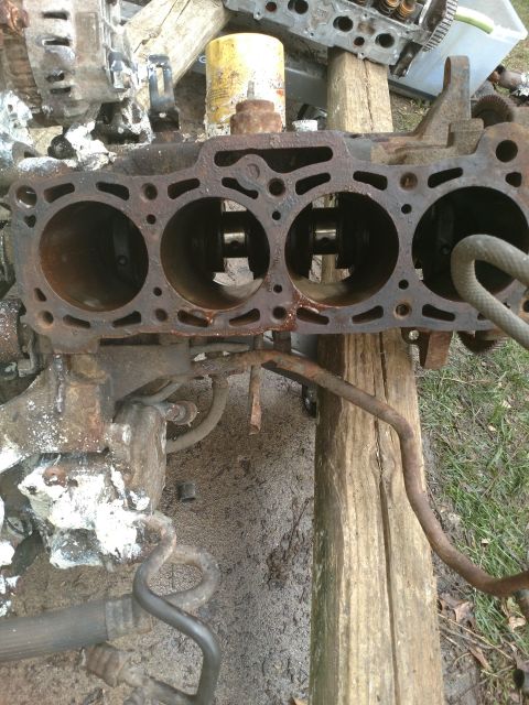 1989 Ford Probe - A heavily rusted engine block with four cylinders is shown outdoors, resting on wooden beams. Various metal parts and hoses are attached to…
