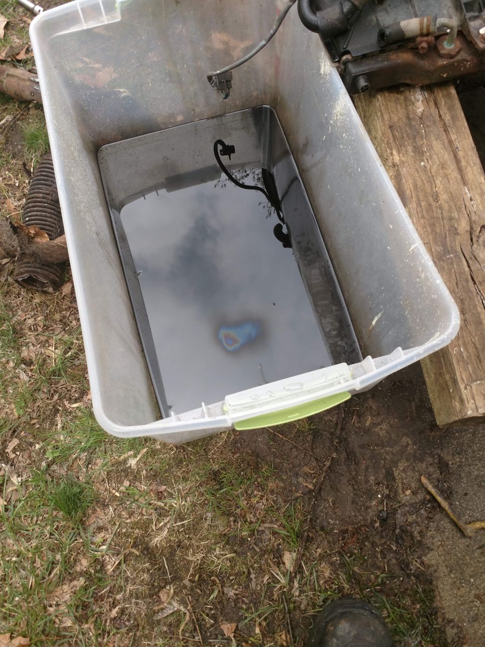 Clear plastic tub with dark liquid and rainbow sheen, black electrical cord submerged.