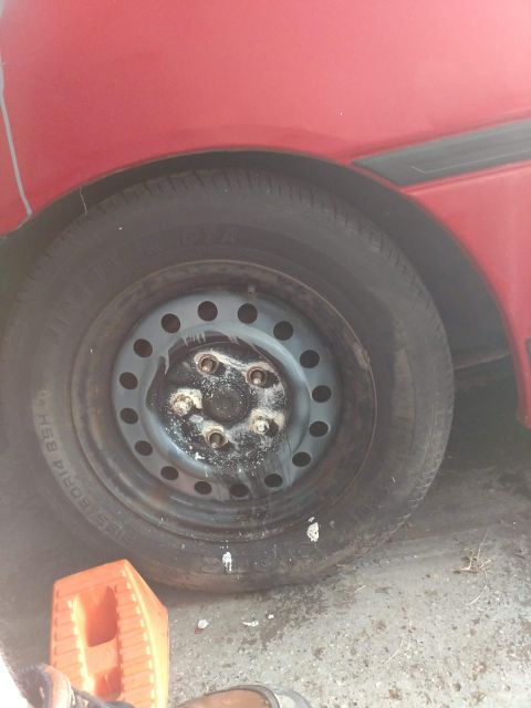 1989 Ford Probe - A close-up view of a red car's wheel and tire, showing a gray steel rim with five lug nuts and white residue. The tire has visible text, an…