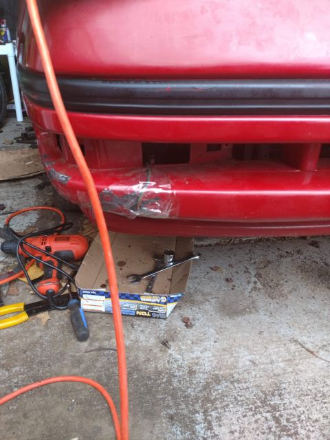 1989 Ford Probe - A close-up view of the front bumper of a red car, showing some damage and repairs made with clear tape. Tools and a box of parts are laid o…