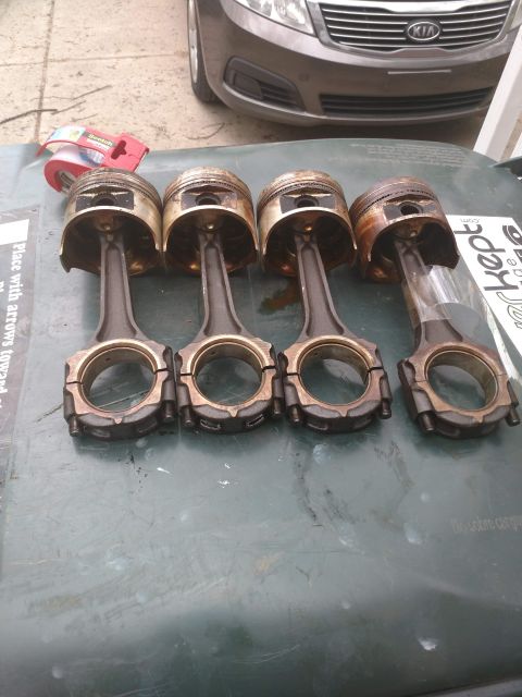 1989 Ford Probe - Four used pistons and connecting rods are laid out on a dark green surface, likely a car's trunk or hood. A roll of Scotch tape and a porti…