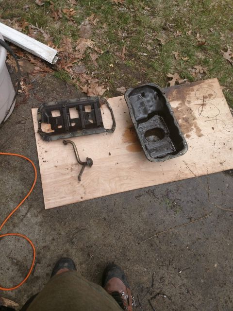 1989 Ford Probe - Two automotive oil pans and an oil pickup tube are laid out on a piece of plywood on the ground. The scene appears to be outdoors with gras…