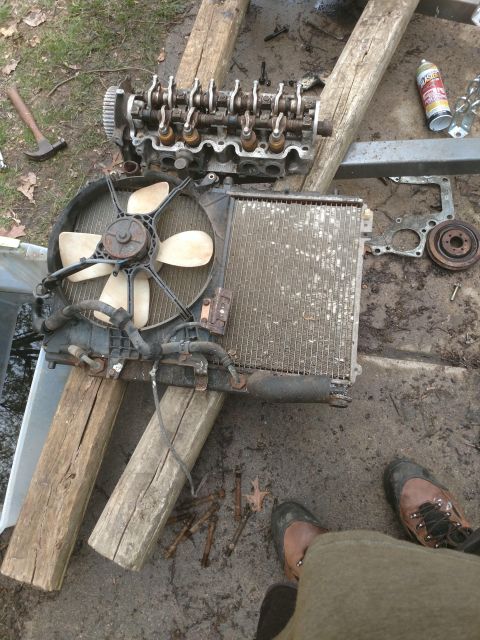 1989 Ford Probe - A disassembled engine and radiator assembly are laid out on wooden beams on the ground. A can of penetrating oil and a hammer are visible n…