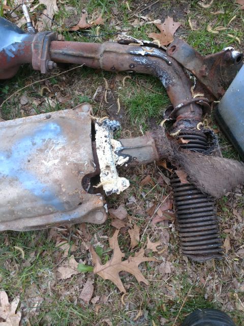 1989 Ford Probe - A rusty exhaust system, possibly from a 1989 Ford Probe, lies on the ground amidst grass and fallen leaves. The system appears damaged with…