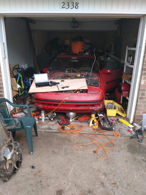 1989 Ford Probe - A red 1989 Ford Probe is partially disassembled in a garage, with its hood open and engine components exposed. Various tools, an orange ext…