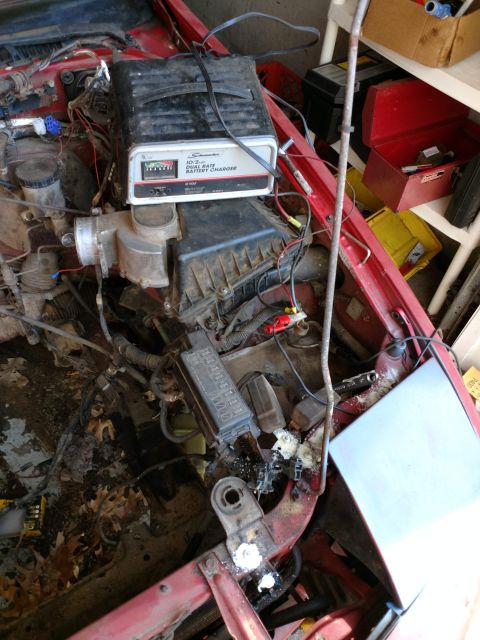 1989 Ford Probe - The engine bay of a red vehicle is shown with a Schumacher battery charger connected to the battery. Various wires, hoses, and a fuse box a…