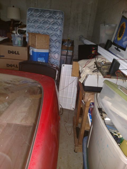 1989 Ford Probe - The rear passenger side of a red 1989 Ford Probe is visible, parked next to a wooden stand and storage bins in a cluttered garage.