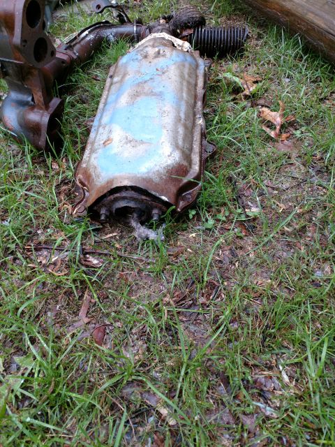 1989 Ford Probe - A rusted catalytic converter and exhaust manifold are lying on the ground amidst grass and debris. The catalytic converter has a light blue…