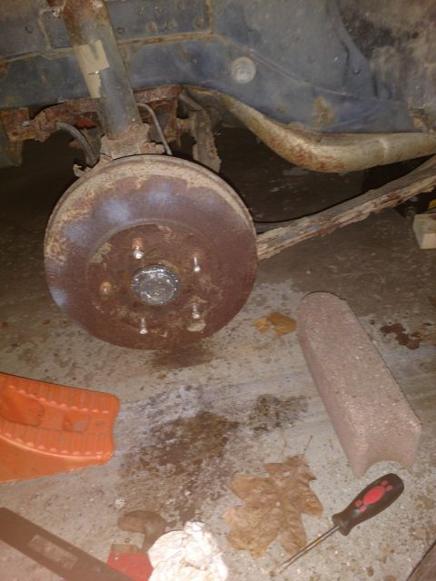 1989 Ford Probe - A rusty brake drum and suspension components are visible on the underside of a vehicle, likely in a garage or driveway setting. Tools and d…