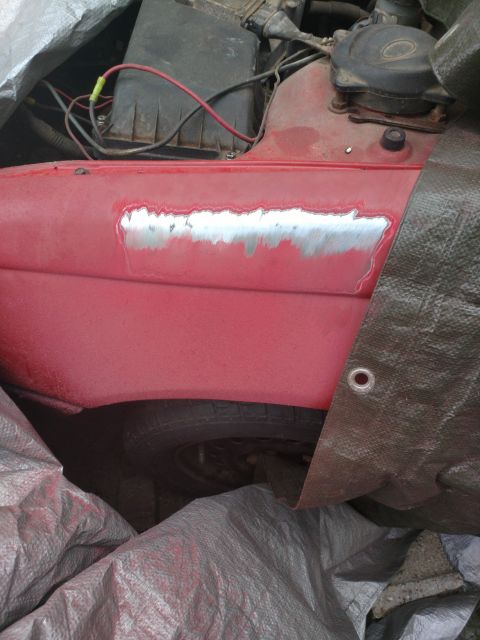 1989 Ford Probe - A close-up view of the driver's side fender of a red car, showing a patch of bare metal where paint has been removed for repair. The vehicl…