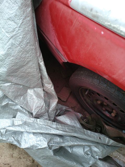 1989 Ford Probe - A close-up view shows the rear fender and tire of a red vehicle partially covered by a silver tarp. The tire is black with a silver rim, an…