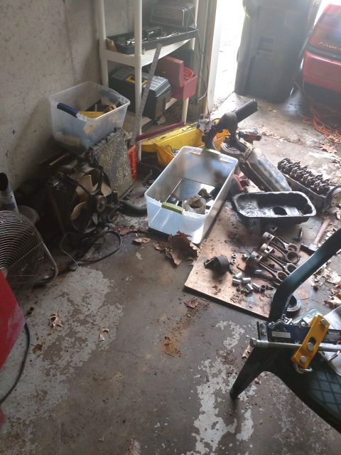 1989 Ford Probe - A cluttered garage scene with various car parts, tools, and storage bins scattered on the concrete floor. A large black trash bin and a red…