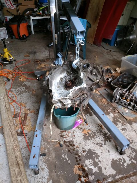 1989 Ford Probe - A disassembled engine and transmission are suspended by an engine hoist in a garage. The engine components are spread out on the concrete f…