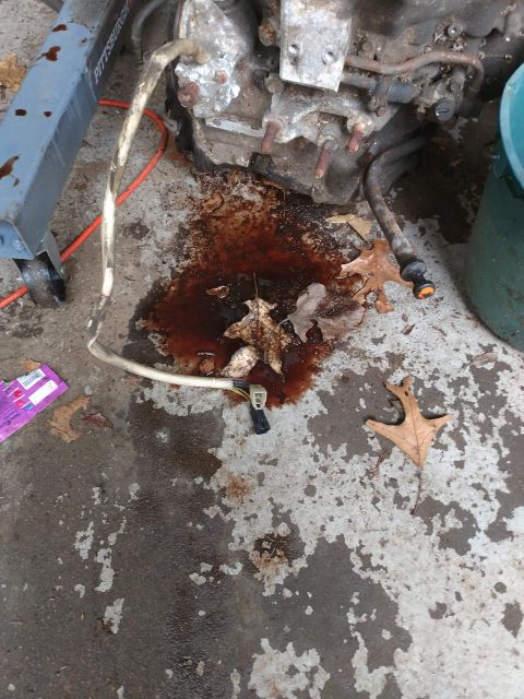 1989 Ford Probe - A dark, oily fluid is leaking onto a concrete surface from a vehicle engine or transmission assembly. A portion of a blue engine hoist is v…