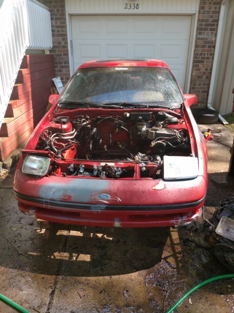 1989 Ford Probe - Front view of a red Ford Probe with its hood open, revealing the engine bay. The car is parked on a wet, muddy driveway in front of a garag…
