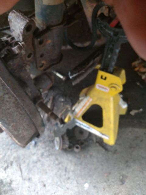 1989 Ford Probe - A yellow jack stand is supporting a vehicle part that appears to be a suspension component. Rusty metal brackets and other unidentifiable p…
