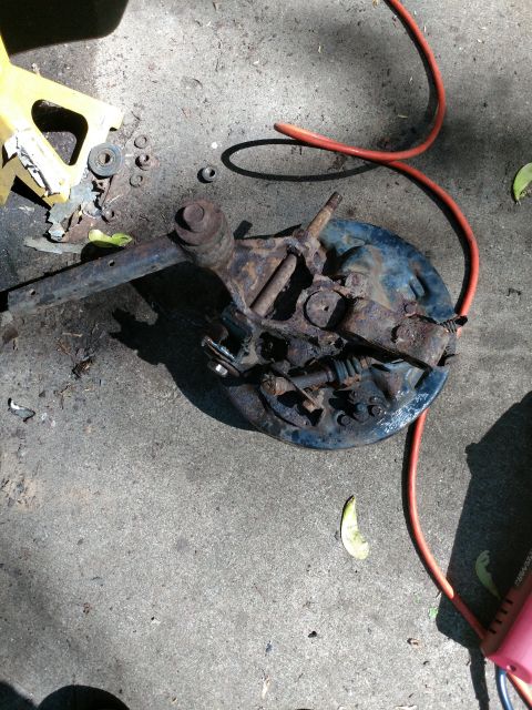 1989 Ford Probe - A heavily rusted brake assembly, likely from a 1989 Ford Probe, lies on a concrete surface next to a yellow jack and an orange cord.