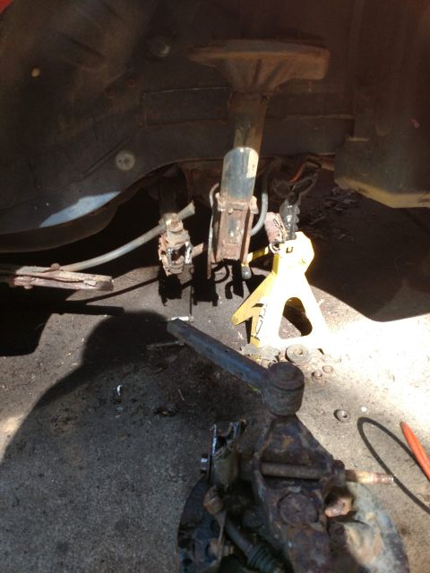 1989 Ford Probe - The underside of a vehicle's suspension system is shown, with a rusty shock absorber, brake components, and a tie rod end. A yellow jack st…