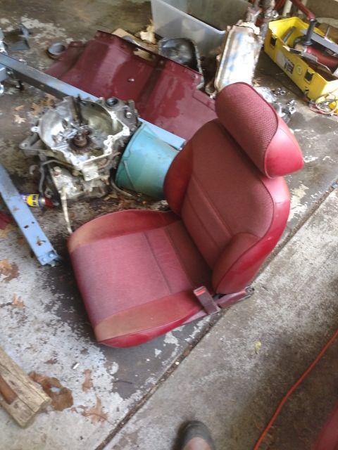1989 Ford Probe - A red car seat with a headrest is shown in a garage setting, surrounded by car parts and tools. A transmission or engine component is visib…