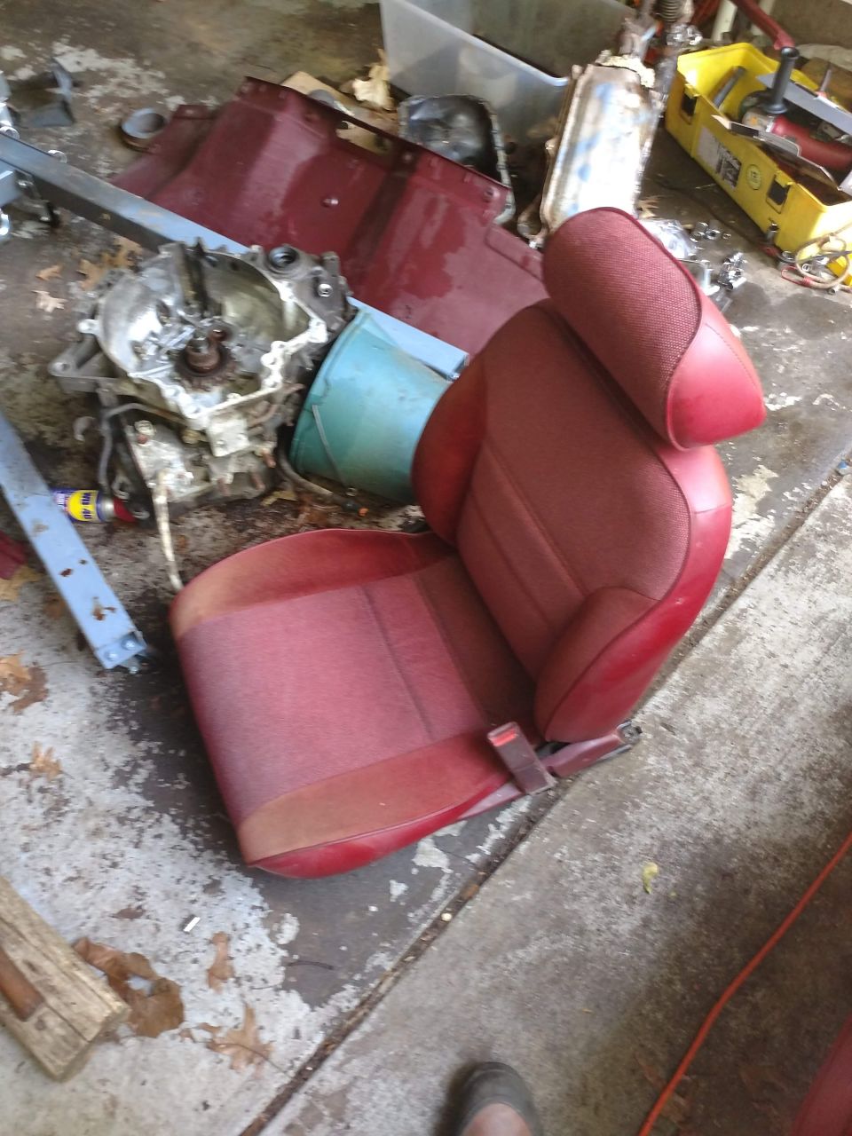 Red car seat with headrest and seatbelt, next to car engine/transmission parts in a garage.