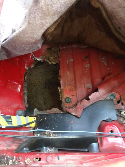 1989 Ford Probe - A large hole in the red painted metal floor of a vehicle reveals significant rust and damage. Wires are visible taped with yellow and gray…