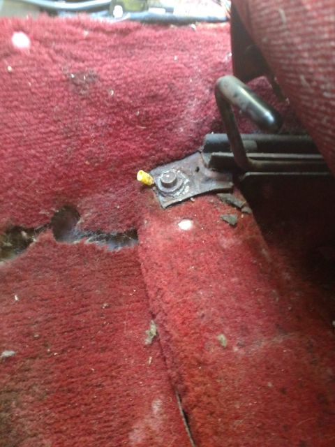 1989 Ford Probe - A close-up view of the rusted metal bracket securing a car seat to the worn red carpet. A small yellow plastic piece is visible near the bo…