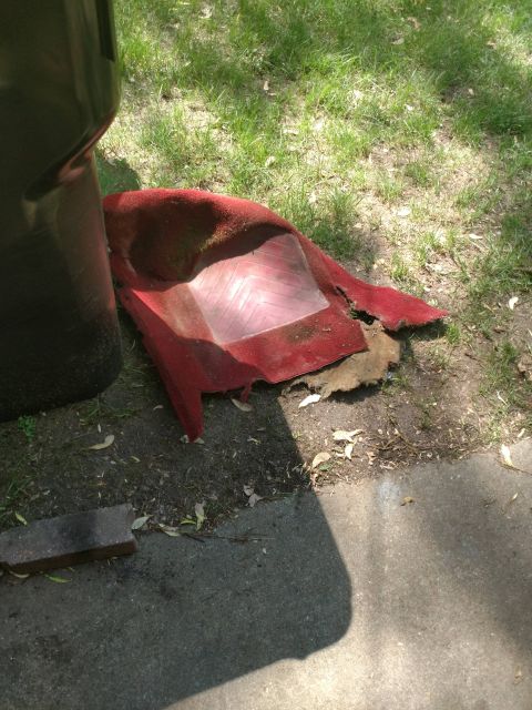 1989 Ford Probe - A red car floor mat is discarded on the grass next to a black trash can. The mat is dirty and torn, with a clear plastic heel protector vis…