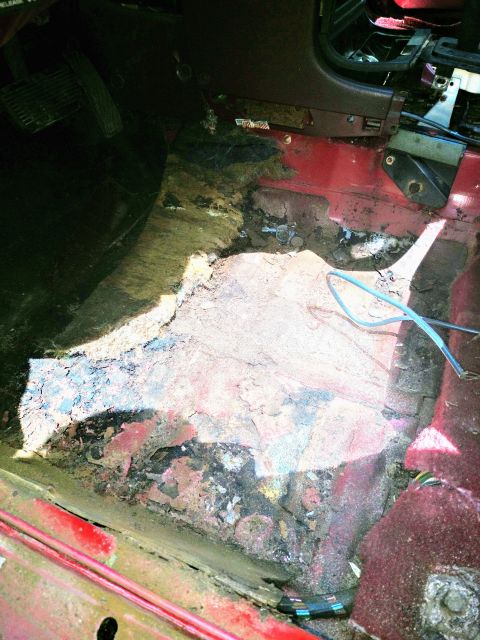 1989 Ford Probe - The interior floor of a vehicle shows significant rust and damage, exposing the red metal underneath and some wiring. The pedals and part o…