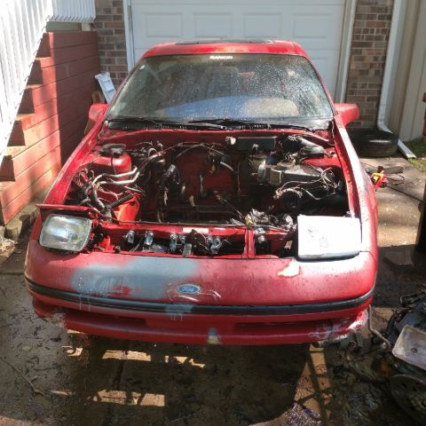 1989 Ford Probe - Front view of a red Ford Probe with its hood open, revealing the engine bay. The car is parked on a wet, dirty surface next to a brick buil…