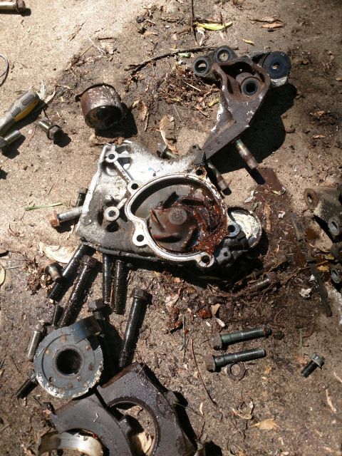 1989 Ford Probe - A disassembled car water pump and associated hardware are laid out on a dirt and concrete surface. The water pump housing is open, revealin…