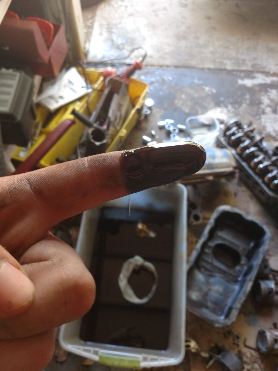 Finger with dark oil residue over a container of oil, with car parts in the background.