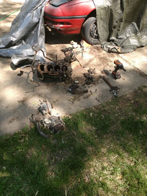 1989 Ford Probe - The engine block and various engine components are disassembled and laid out on the ground next to a red car covered by a tarp. A cordless…