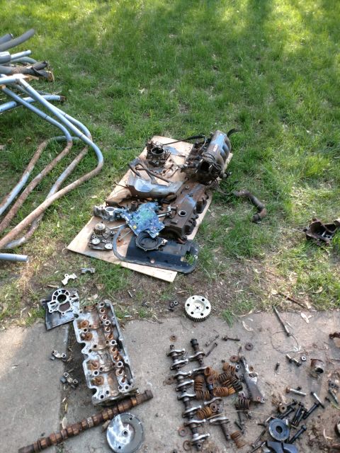 1989 Ford Probe - Disassembled engine parts are laid out on a grassy area and concrete. Visible components include a cylinder head, camshaft, valves, springs…