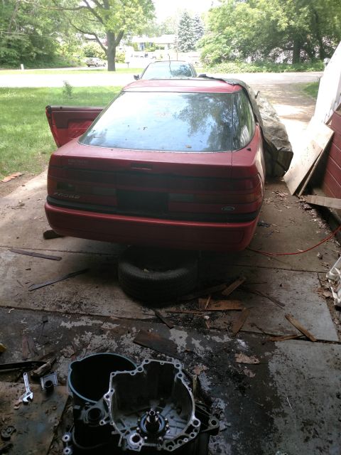 1989 Ford Probe - The rear of a red Ford Probe is shown, jacked up on a tire with its driver's side door open. A disassembled transmission and tools are visi…