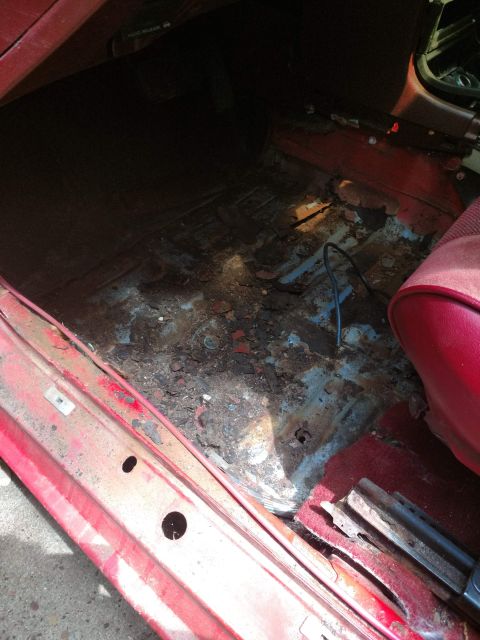 1989 Ford Probe - The interior floor of a red vehicle shows significant rust and damage, exposing the underlying metal structure. A black wire is visible on…