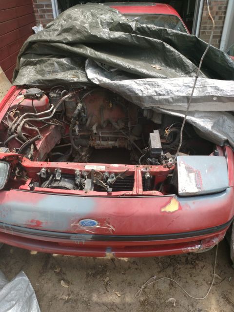 1989 Ford Probe - The front of a red Ford Probe is partially covered by a large, dirty tarp. The engine bay is exposed, showing various hoses and components,…