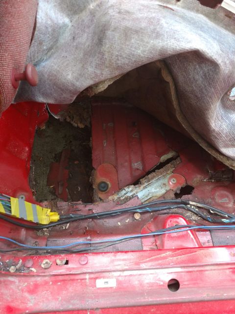 1989 Ford Probe - The interior floor of a red vehicle shows significant rust and damage, with exposed wiring and torn upholstery.