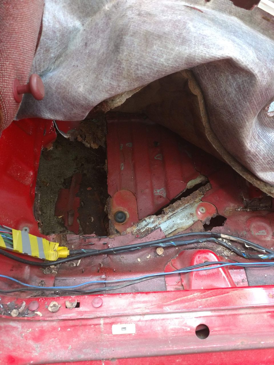 Rusted out interior floor of a red car with exposed wiring and torn fabric.
