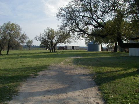 1991 Ford Bronco - Gravel ranch lane running through a green pasture toward a metal barn, a corrugated grain bin and a white outbuilding, with live oaks shadi…