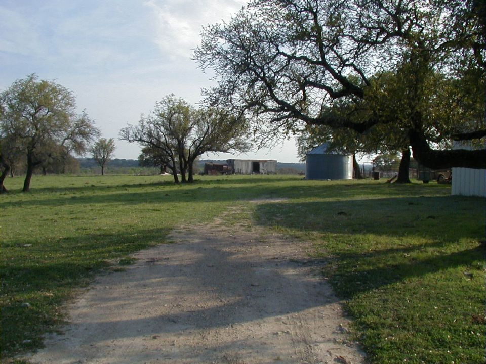 Gravel ranch lane leading to a metal barn and grain bin under live oaks