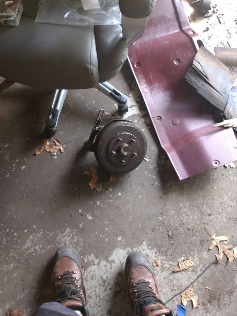 1989 Ford Probe - A close-up, top-down view of a rusty brake drum assembly with a metal bracket attached, resting on a concrete floor next to a maroon-colore…