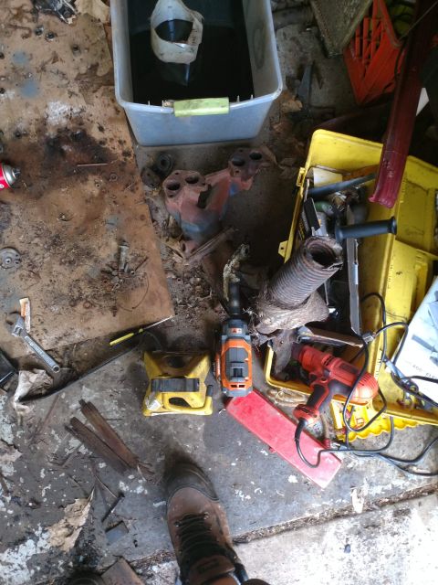 1989 Ford Probe - A collection of automotive parts and tools are scattered on a dirty concrete floor. Items include an exhaust manifold, a yellow jack, a cor…