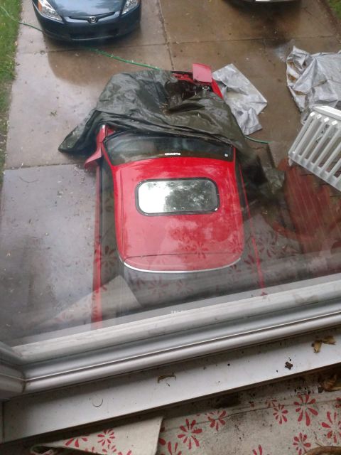 1989 Ford Probe - A red convertible sports car is partially covered by a dark tarp in a driveway. Another car is visible in the background, and the scene app…