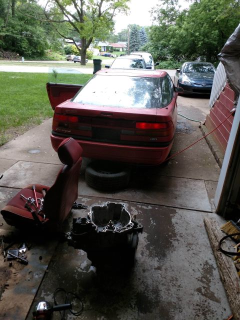 1989 Ford Probe - A red Ford Probe is parked in a driveway with its driver's side door open. A car seat and transmission are on the ground next to the car.