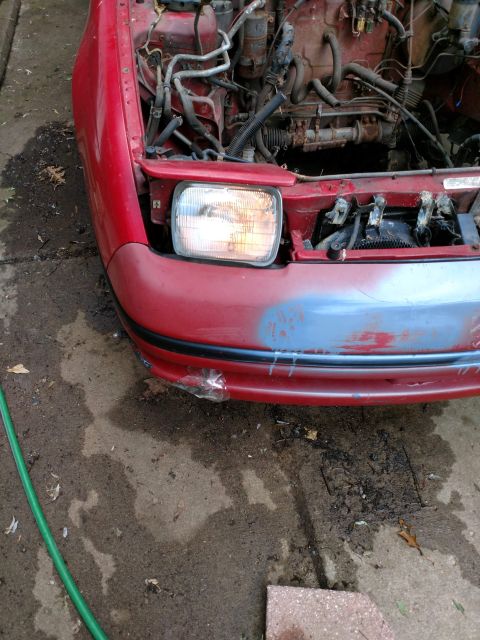 1989 Ford Probe - A red 1989 Ford Probe is shown from the front with its hood open, revealing the engine bay. The front bumper has been spray-painted with a…