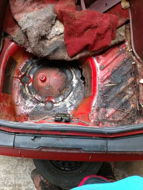 1989 Ford Probe - The interior of the trunk of a red Ford Probe shows significant rust and damage to the floor pan, with some red and gray carpet remnants vi…