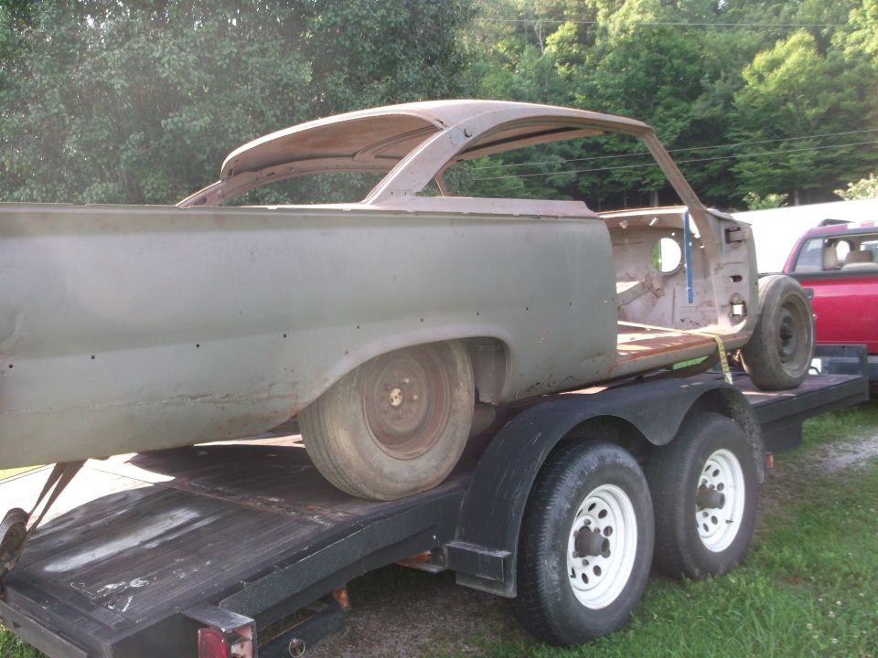 Gray car body on a trailer, missing doors and roof, with a red truck visible behind it
